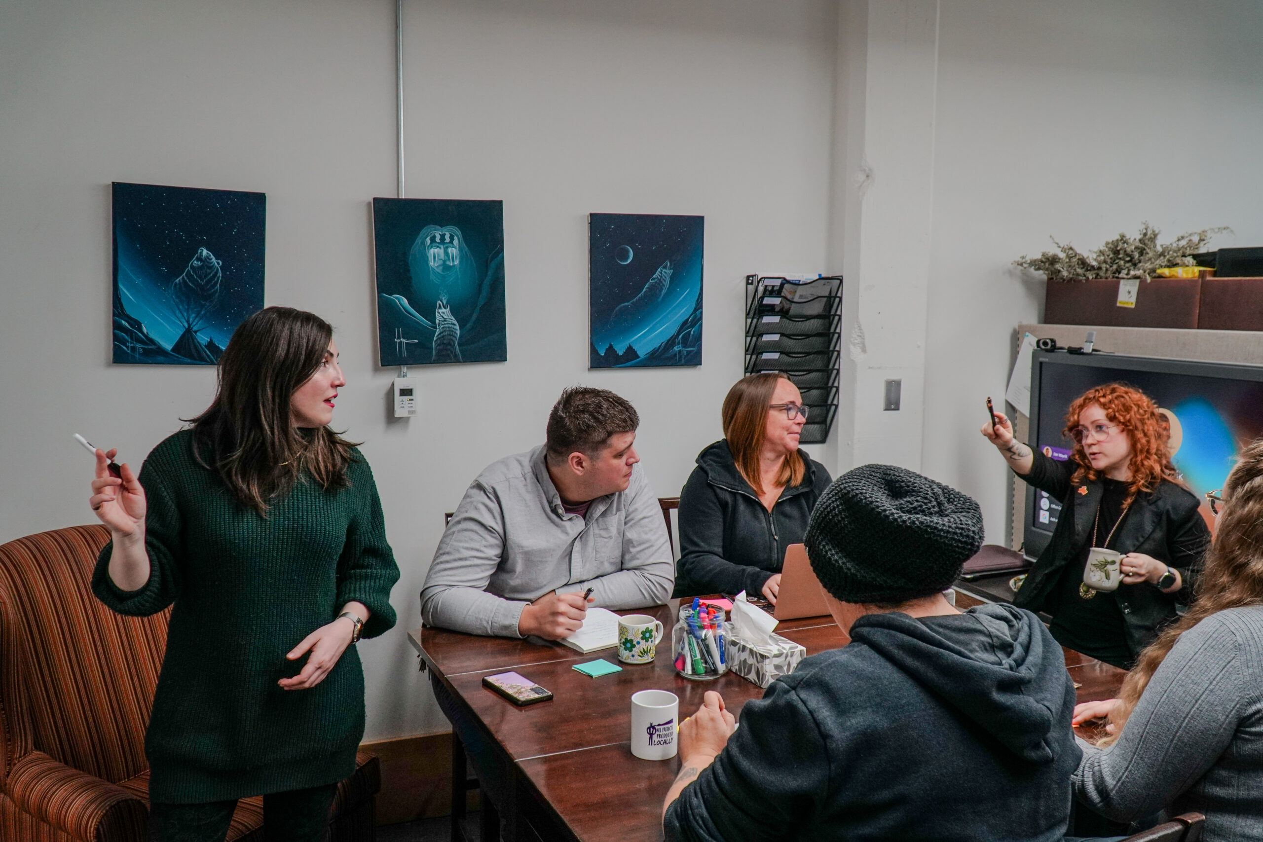A group of people sit and stand around a meeting table during a collaborative discussion. Various supplies, mugs, and notebooks are on the table.